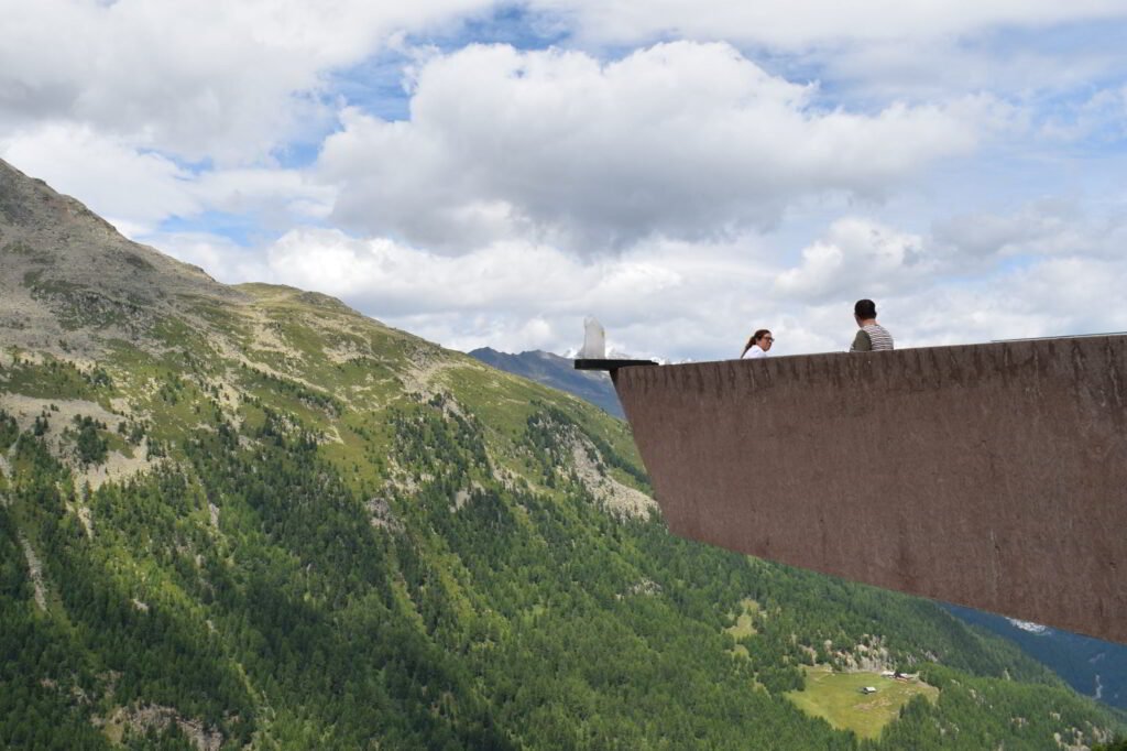 Kaksi ihmistä Timmelsjoch alppitien Walkaway-näköalatasanteella. Taustalla upeat Alpit.